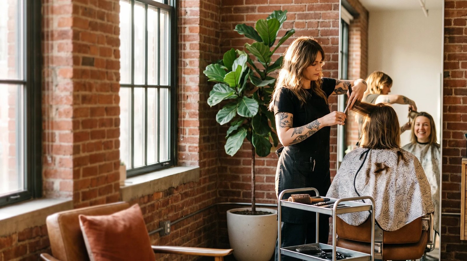A tattooed stylist trimming a client's long brown hair in a sunlit brick-walled salon beside a fiddle-leaf fig, while the smiling client watches in the mirror