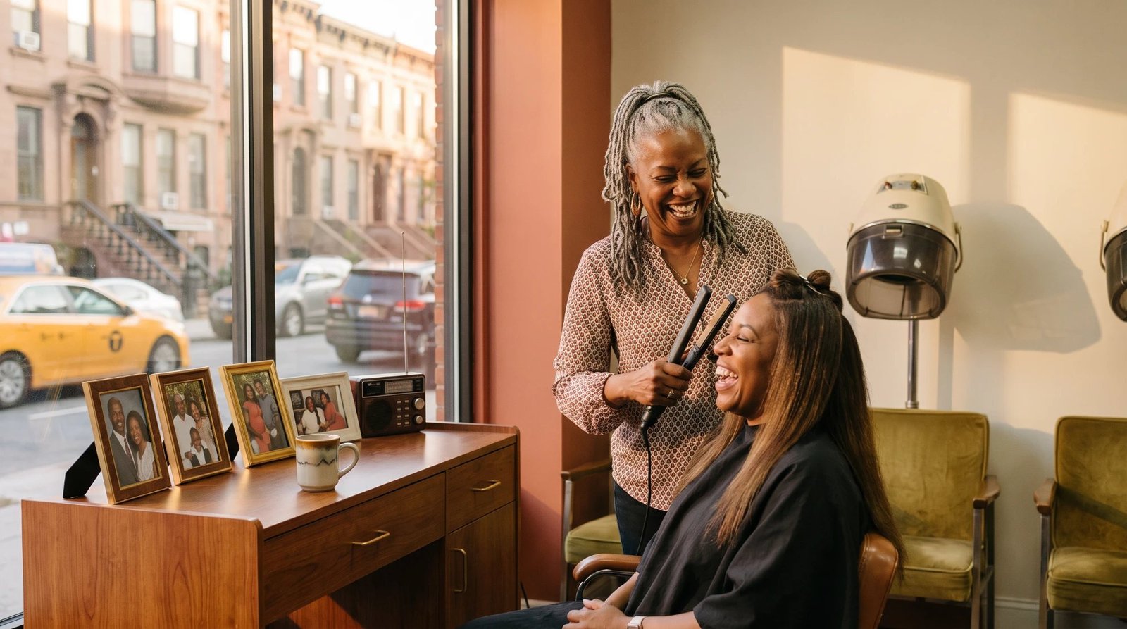 A stylist with gray locs laughing with a client as she flat-irons her long hair beside the salon window, with brownstones and a yellow taxi on the street outside